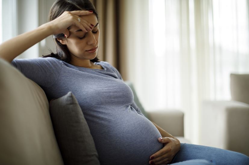 Une femme enceinte portant un t-shirt bleu est assise sur un canapé clair dans un salon lumineux, une main posée sur son front en signe de fatigue et l’autre sur son ventre arrondi.