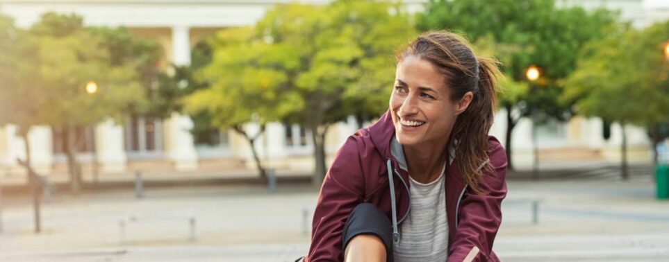 Femme souriante assise sur un ponton en bois, lace ses baskets de running gris et bleu, vêtue d’un coupe-vent bordeaux et d’un legging noir, smartphone et écouteurs posés à côté.