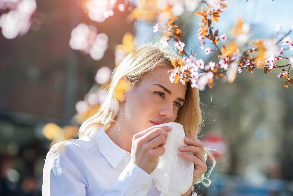 Femme en extérieur sous des branches fleuries, se couvrant le nez avec un mouchoir à cause d’une allergie printanière