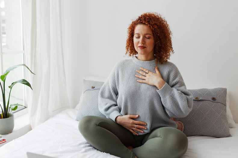 Femme enceinte rousse assise en tailleur sur un lit blanc, les yeux fermés, main droite sur la poitrine et main gauche sur le ventre, pratiquant une respiration consciente dans une chambre lumineuse avec coussins et plante à proximité.