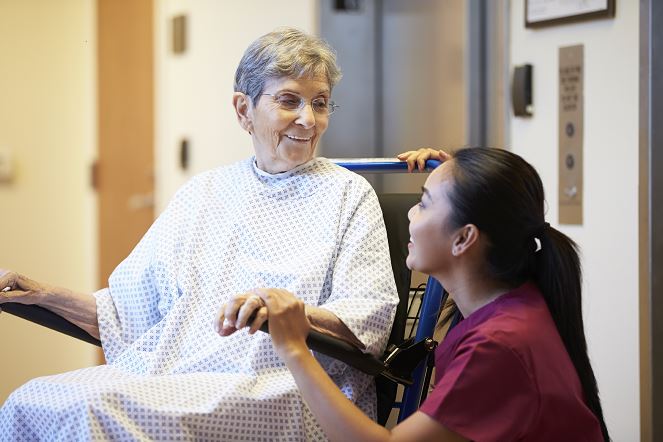 Une patiente âgée en blouse d’hôpital est assise dans un fauteuil roulant et sourit à une infirmière en uniforme bordeaux, agenouillée à côté d’elle dans un couloir médical et lui tenant la main.