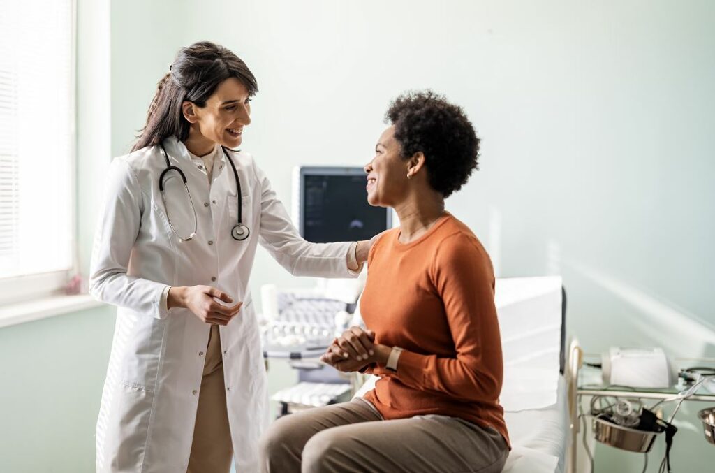 Femme médecin en blouse blanche souriante, stéthoscope autour du cou, posant la main sur l'épaule d'une patiente noire assise sur une table d'examen, avec un appareil d'échographie en arrière-plan