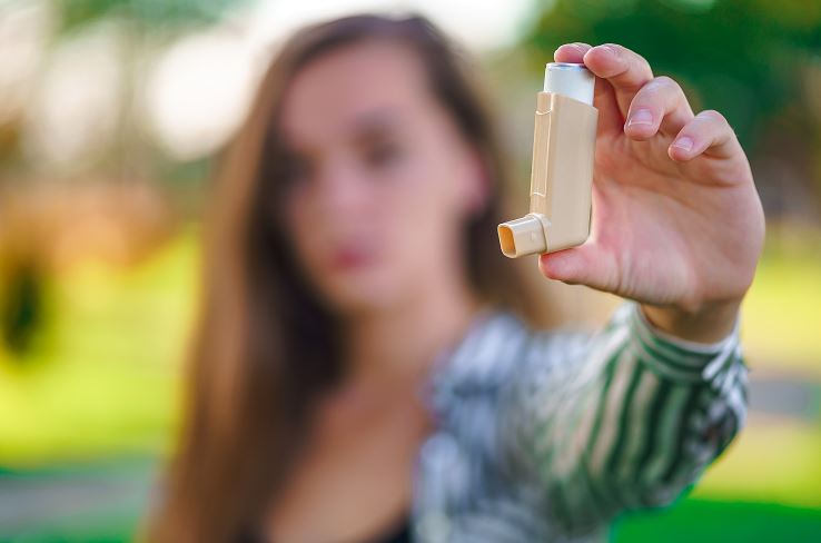 Gros plan sur une femme tenant un inhalateur d'asthme beige dans un parc au décor flou, illustrant un traitement respiratoire mobile