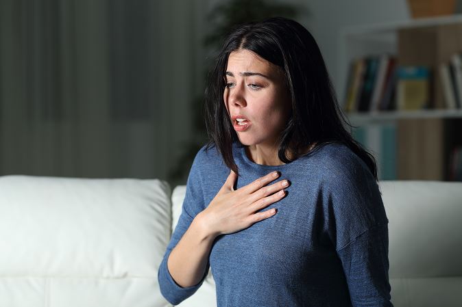 Femme assise sur un canapé dans un salon peu éclairé, main posée sur la poitrine, regard inquiet et bouche entrouverte, illustrant une crise d’angoisse nocturne