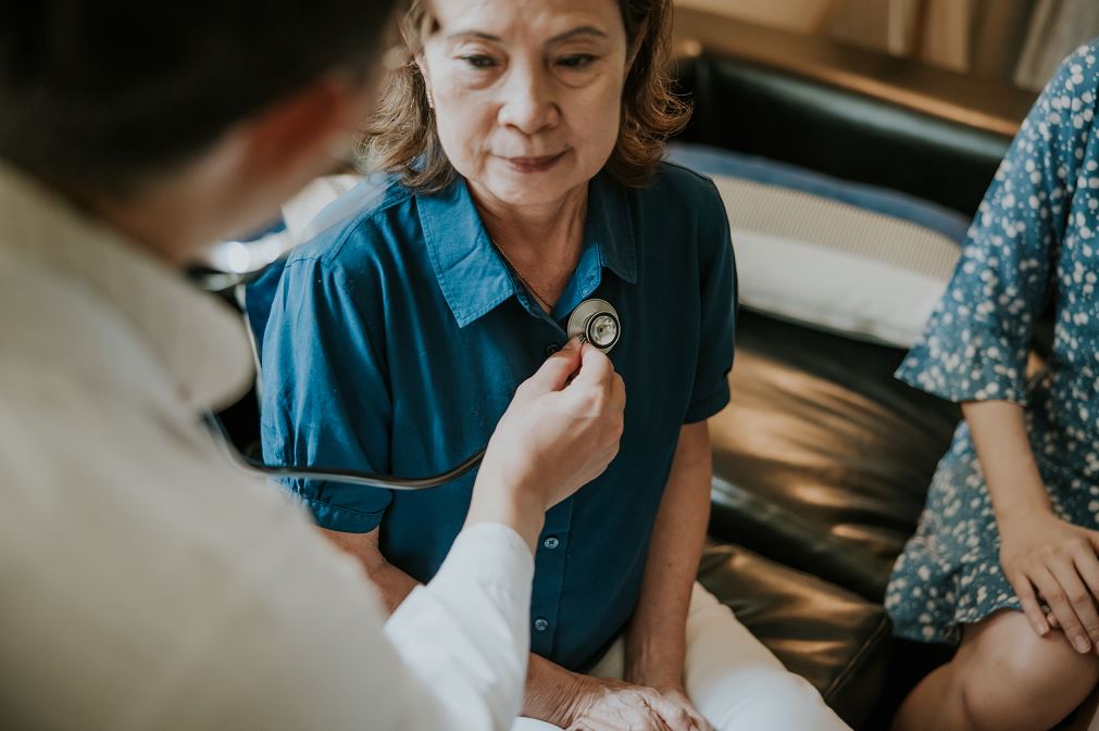 Un médecin ausculte la poitrine d’une patiente âgée en visite médicale à domicile, assise sur un canapé près d’un accompagnant.