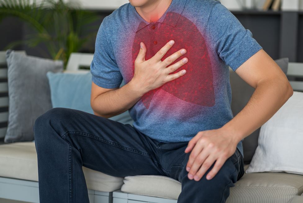 Homme assis sur un canapé, main posée sur la poitrine, visage crispé, superposition rouge en forme de poumon illustrant une douleur thoracique évoquant un problème cardiaque ou pulmonaire.