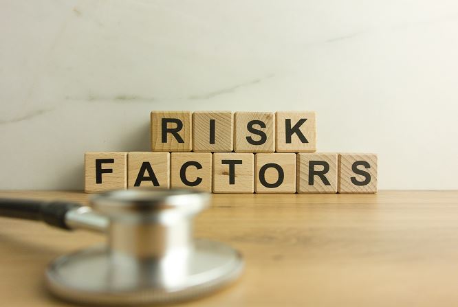 Photo de blocs de bois formant le texte 'RISK FACTORS' posés sur une surface en bois, avec au premier plan un stéthoscope médical flou, illustrant l’évaluation des facteurs de risque en santé.