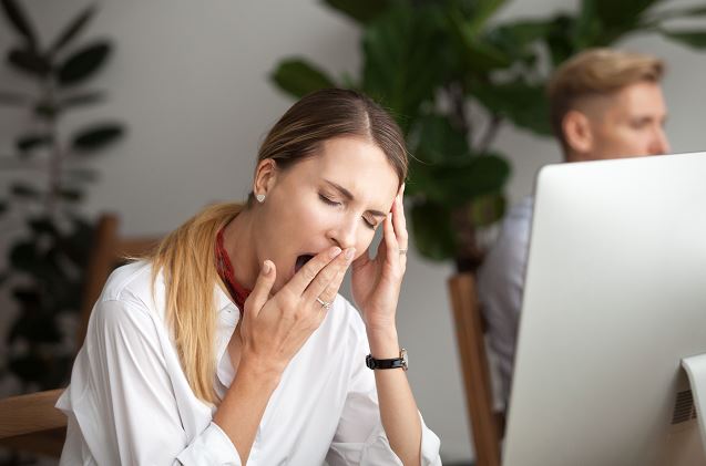 Jeune femme assise devant son ordinateur au bureau, bâillant et se tenant la tête pour soulager la fatigue. En arrière-plan, un collègue flou et une plante verte décorent l’espace de travail.