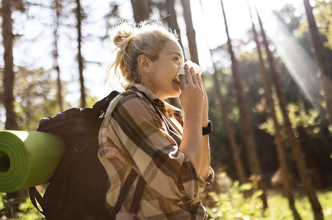 Jeune femme blonde en chemise à carreaux et sac à dos avec tapis de sol vert, se mouche dans un mouchoir en plein milieu d’une forêt baignée de lumière et d’arbres hauts