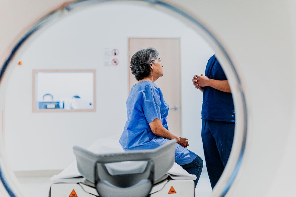 Patiente senior assise sur la table d’un scanner corporel, en conversation avec un soignant visible de profil, vue à travers l’anneau de l’appareil dans une salle d’imagerie médicale claire.