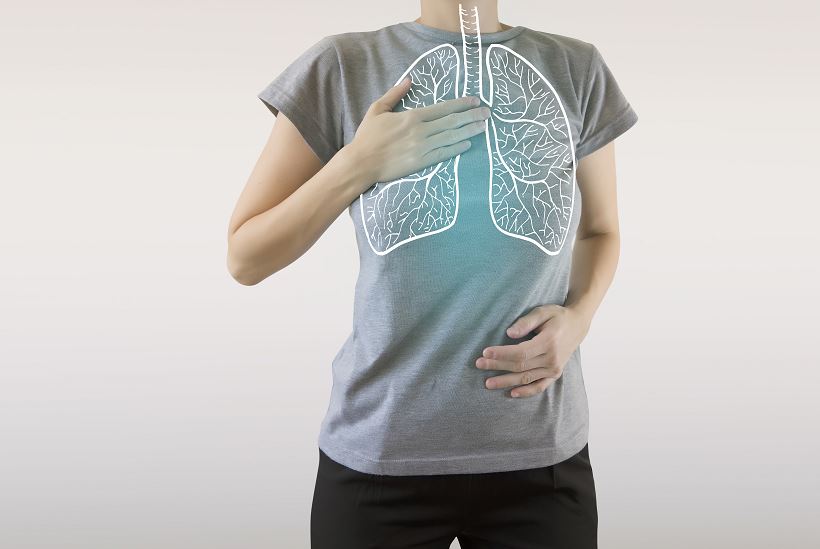 Personne debout de face, main droite posée sur la poitrine et main gauche sur l’abdomen, portant un t-shirt gris sur lequel est superposé un schéma blanc de l’anatomie des poumons pour illustrer la santé respiratoire.