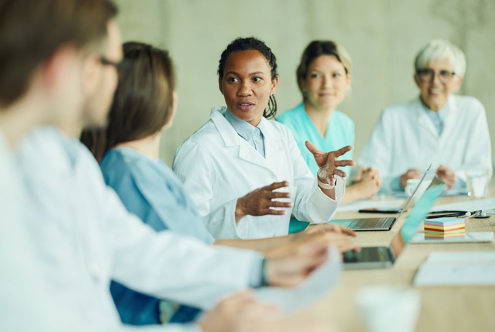 Médecin afro-descendante en blouse blanche gesticulant lors d'une réunion avec collègues médecins et infirmières autour d'une table avec ordinateurs portables.