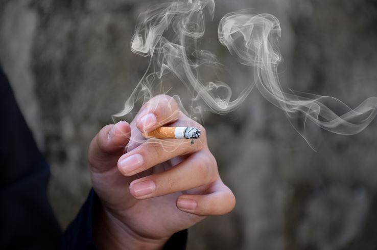 Gros plan sur une main tenant une cigarette allumée, la fumée tourbillonnant en volutes grises contre un mur flou en arrière-plan