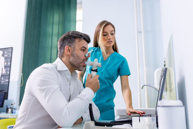 Homme assis réalisant un test de spirométrie avec un masque nasal relié à un spiromètre sous la supervision d’une infirmière en blouse bleue dans un cabinet médical