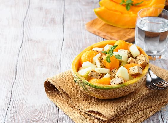 Demi-melon évidé servant de saladier, garni de billes de melon orange, dés de feta assaisonnés d’herbes fraîches et poivre, posé sur un napperon en jute clair à côté d’un verre d’eau et de deux fourchettes.