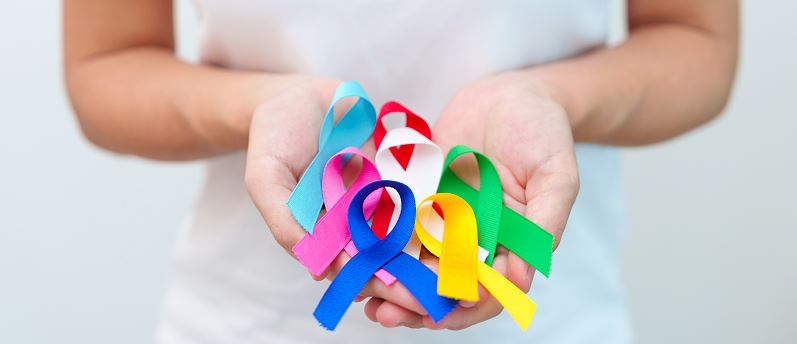 Gros plan sur deux mains ouvertes tenant plusieurs rubans de sensibilisation multicolores (turquoise, rose, bleu, jaune, vert et rouge) symbolisant différentes causes de santé et sociales.